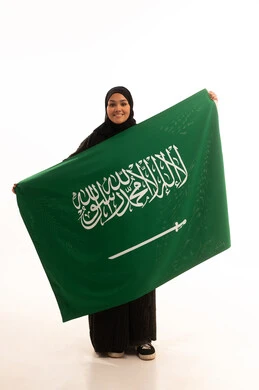 ذكرى اليوم الوطني السعودي 23 سبتمبر، يوم العلم 11 مارس، مفهوم حب الوطن والانتماء اليه، بورتريه لسيدة عربية خليجية سعودية محجبة مبتسمة ترتدي عباءة سوداء تحمل بيدها علم المملكة وتنظر الى الكاميرا بإيماءات السرور والثقة بالنفس، صورة الجسم بالكامل، خلفية بيضاء