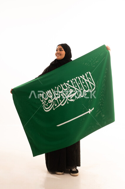الوقوف باستقامة بايماءات الاعتزاز والفخربالانتماء إلى الوطن، ذكرى اليوم الوطني السعودي 23 سبتمبر، يوم العلم 11 مارس، إظهار الحب والولاء للبلاد، بورتريه لسيدة عربية خليجية سعودية محجبة ترتدي عباءة سوداء تحمل بيدها علم المملكة، صورة الجسم بالكامل، خلفية بيضاء