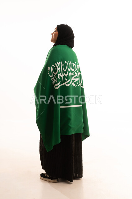 الحب والولاء للبلاد، التطلع والنظر إلى الاعلى بايماءات الاعتزاز والفخربالانتماء إلى الوطن، اليوم الوطني السعودي 23 سبتمبر، يوم العلم 11 مارس، بورتريه من الخلف لسيدة عربية خليجية سعودية محجبة ترتدي عباءة تضع على كتفيهاعلم المملكة، صورة الجسم بالكامل، خلفية بيضاء