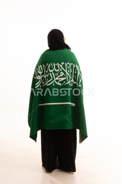 الحب والولاء للبلاد، التطلع والنظر إلى الاعلى بايماءات الاعتزاز والفخربالانتماء إلى الوطن، اليوم الوطني السعودي 23 سبتمبر، يوم العلم 11 مارس، بورتريه من الخلف لسيدة عربية خليجية سعودية محجبة ترتدي عباءة تضع على كتفيهاعلم المملكة، صورة الجسم بالكامل، خلفية بيضاء