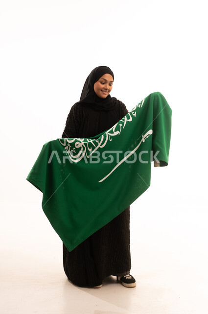 النظر إلى العلم بايماءات الاعتزاز والفخربالانتماء إلى الوطن، ذكرى اليوم الوطني السعودي 23 سبتمبر، يوم العلم 11 مارس، إظهار الحب والولاء للبلاد، بورتريه لسيدة عربية خليجية سعودية محجبة ترتدي عباءة سوداء تحمل بيدها علم المملكة، صورة الجسم بالكامل، خلفية بيضاء
