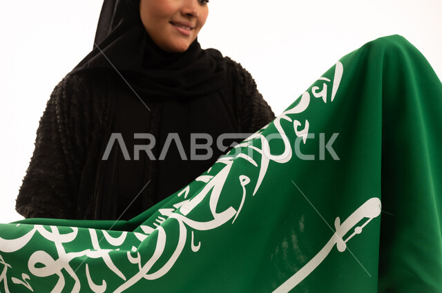 راية التوحيد والإسلام، إحياء ذكري اليوم الوطني السعودي 23 سبتمبر، الفخر والاعتزاز بالانتماء للبلاد، يوم العلم 11 مارس، مفهوم حب الوطن، بورتريه مقرب لسيدة عربية خليجية سعودية محجبة مبتسمة ترتدي عباءة سوداء تحمل بيدها علم المملكة، خلفية بيضاء