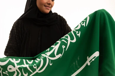 Banner of monotheism and Islam, commemorating Saudi National Day September 23, pride and honor of belonging to the country, Flag Day March 11, concept of patriotism, close-up portrait of a smiling veiled Saudi Arabian Gulf woman wearing a black abaya holding the flag of the Kingdom, white background