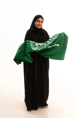 Flag Day, March 11, the concept of patriotism and belonging to the homeland, the memory of Saudi National Day, September 23, a portrait of a smiling veiled Saudi Arabian Gulf woman wearing a black abaya, holding the flag of the Kingdom in her hand and looking at the camera with gestures of pleasure and self-confidence, a full body image, a white background