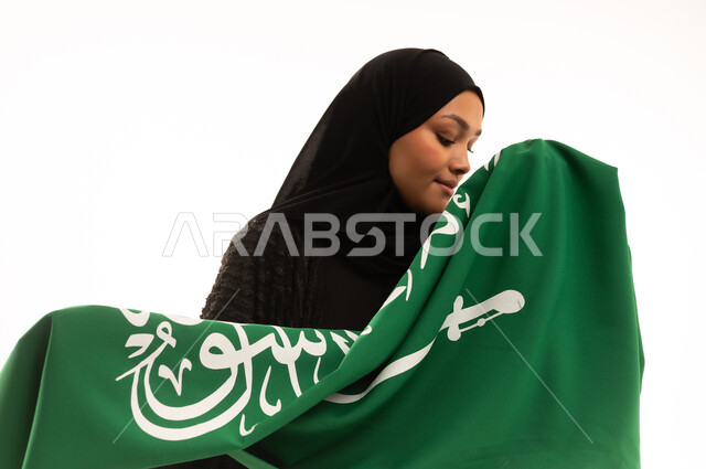 Banner of monotheism and Islam, commemorating Saudi National Day September 23, pride and honor of belonging to the country, Flag Day March 11, concept of patriotism, close-up portrait of a smiling veiled Saudi Arabian Gulf woman wearing a black abaya holding the flag of the Kingdom, white background