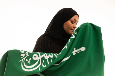 Banner of monotheism and Islam, commemorating Saudi National Day September 23, pride and honor of belonging to the country, Flag Day March 11, concept of patriotism, close-up portrait of a smiling veiled Saudi Arabian Gulf woman wearing a black abaya holding the flag of the Kingdom, white background