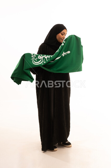 رفع راية التوحيد والإسلام، إحياء ذكري اليوم الوطني السعودي 23 سبتمبر، الفخر والاعتزاز بالانتماء للبلاد، يوم العلم 11 مارس، مفهوم حب الوطن، بورتريه لسيدة عربية خليجية سعودية محجبة مبتسمة ترتدي عباءة سوداء تحمل بيدها علم المملكة، الجسم بالطول الكامل، خلفية بيضاء