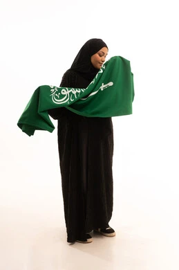 رفع راية التوحيد والإسلام، إحياء ذكري اليوم الوطني السعودي 23 سبتمبر، الفخر والاعتزاز بالانتماء للبلاد، يوم العلم 11 مارس، مفهوم حب الوطن، بورتريه لسيدة عربية خليجية سعودية محجبة مبتسمة ترتدي عباءة سوداء تحمل بيدها علم المملكة، الجسم بالطول الكامل، خلفية بيضاء