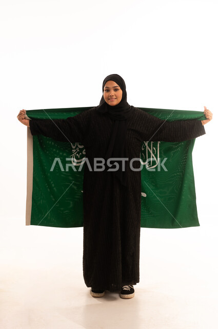 ذكرى اليوم الوطني السعودي 23 سبتمبر، يوم العلم 11 مارس، مفهوم حب الوطن والانتماء اليه، بورتريه لسيدة عربية خليجية سعودية محجبة مبتسمة ترتدي عباءة سوداء تحمل بيدها علم المملكة وتنظر الى الكاميرا بإيماءات السرور والثقة بالنفس، صورة الجسم بالكامل، خلفية بيضاء