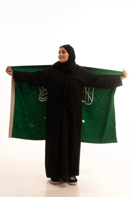 الوقوف باستقامة بايماءات الاعتزاز والفخربالانتماء إلى الوطن، ذكرى اليوم الوطني السعودي 23 سبتمبر، يوم العلم 11 مارس، إظهار الحب والولاء للبلاد، بورتريه لسيدة عربية خليجية سعودية محجبة ترتدي عباءة سوداء تحمل بيدها علم المملكة، صورة الجسم بالكامل، خلفية بيضاء