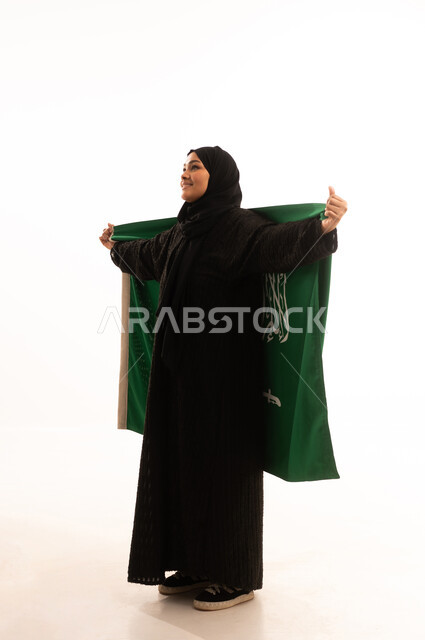 التطلع والنظر إلى الاعلى بايماءات الاعتزاز والفخربالانتماء إلى الوطن، ذكرى اليوم الوطني السعودي 23 سبتمبر، يوم العلم 11 مارس، إظهار الحب والولاء للبلاد، بورتريه لسيدة عربية خليجية سعودية محجبة ترتدي عباءة تحمل بيدها علم المملكة، صورة الجسم بالكامل، خلفية بيضاء