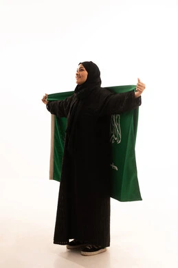 التطلع والنظر إلى الاعلى بايماءات الاعتزاز والفخربالانتماء إلى الوطن، ذكرى اليوم الوطني السعودي 23 سبتمبر، يوم العلم 11 مارس، إظهار الحب والولاء للبلاد، بورتريه لسيدة عربية خليجية سعودية محجبة ترتدي عباءة تحمل بيدها علم المملكة، صورة الجسم بالكامل، خلفية بيضاء