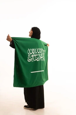 الحب والولاء للبلاد، التطلع والنظر إلى الاعلى بايماءات الاعتزاز والفخربالانتماء إلى الوطن، اليوم الوطني السعودي 23 سبتمبر، يوم العلم 11 مارس، بورتريه من الخلف لسيدة عربية خليجية سعودية محجبة ترتدي عباءة تضع على كتفيهاعلم المملكة، صورة الجسم بالكامل، خلفية بيضاء
