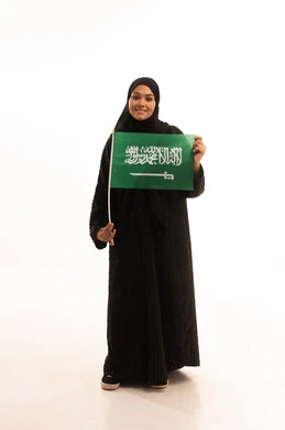 يوم العلم 11 مارس، مفهوم حب الوطن والانتماء اليه، ذكرى اليوم الوطني السعودي 23 سبتمبر، بورتريه لسيدة عربية خليجية سعودية محجبة مبتسمة ترتدي عباءة سوداء تحمل بيدها علم المملكة وتنظر الى الكاميرا بإيماءات السرور والثقة بالنفس، صورة الجسم بالكامل، خلفية بيضاء