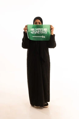 Saudi National Day commemoration, September 23, Flag Day, March 11, the concept of love and belonging to one's homeland, a portrait of a smiling, veiled Saudi Arabian Gulf woman wearing a black abaya, holding the Kingdom's flag in her hand and looking at the camera with gestures of pleasure and self-confidence, a full body image, a white background