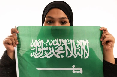 Pride and pride in belonging to the country, Saudi National Day September 23, Flag Day March 11, concept of patriotism, close-up portrait of a smiling veiled Saudi Arabian Gulf woman wearing a black abaya holding the Kingdom's flag in her hand and looking at the camera with gestures of self-confidence, white background