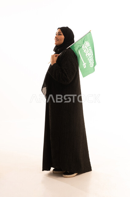 الوقوف باستقامة بايماءات الاعتزاز والفخربالانتماء إلى الوطن، ذكرى اليوم الوطني السعودي 23 سبتمبر، يوم العلم 11 مارس، إظهار الحب والولاء للبلاد، بورتريه لسيدة عربية خليجية سعودية محجبة ترتدي عباءة سوداء تحمل بيدها علم المملكة، صورة الجسم بالكامل، خلفية بيضاء