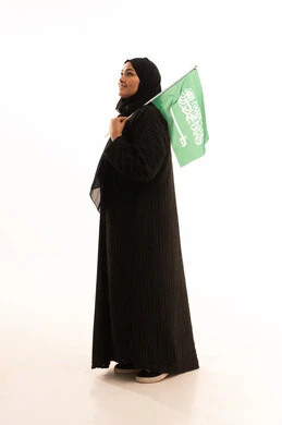 Standing up straight with gestures of pride and belonging to the homeland, Saudi National Day, September 23, Flag Day, March 11, showing love and loyalty to the country, portrait of a veiled Saudi Arabian Gulf woman wearing a black abaya and holding the flag of the Kingdom, full body image, white background