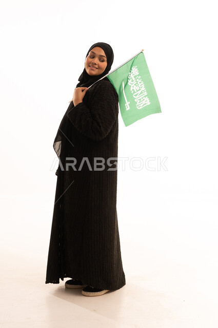 النظر إلى العلم بايماءات الاعتزاز والفخربالانتماء إلى الوطن، ذكرى اليوم الوطني السعودي 23 سبتمبر، يوم العلم 11 مارس، إظهار الحب والولاء للبلاد، بورتريه لسيدة عربية خليجية سعودية محجبة ترتدي عباءة سوداء تحمل بيدها علم المملكة، صورة الجسم بالكامل، خلفية بيضاء