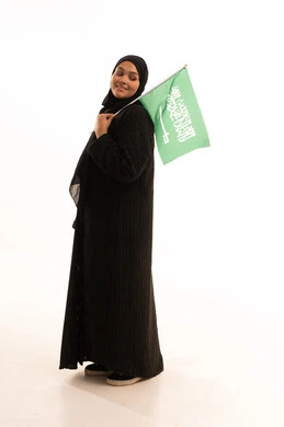 Looking at the flag with gestures of pride and belonging to the homeland, Saudi National Day, September 23, Flag Day, March 11, showing love and loyalty to the country, portrait of a veiled Saudi Arabian Gulf woman wearing a black abaya holding the flag of the Kingdom, full body image, white background