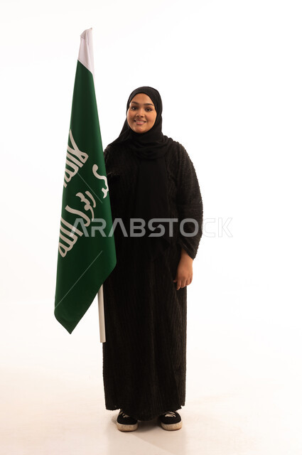 النظر إلى الكاميرا بإيماءات الاعتزاز والشموخ، ذكرى تأسيس الدولة السعودية الأولى 1727م، الفخر بإنجازات المملكة، يوم بدينا 22 فبراير، إظهار الولاء والحب للوطن, بورتريه لامرأة عربية خليجية سعودية ترتدي العباءة تحمل علم التأسيس، الجسم بالطول الكامل، خلفية بيضاء