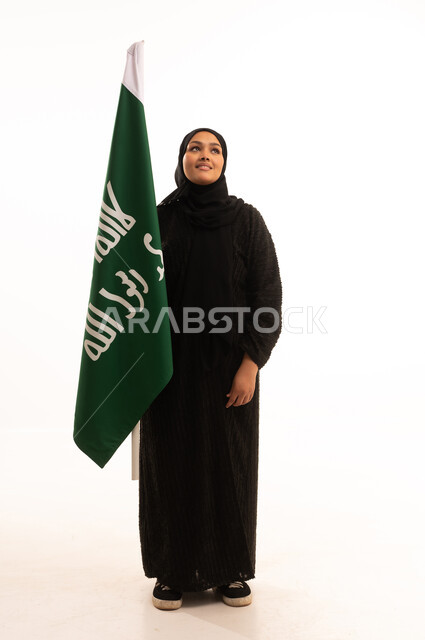 ذكرى تأسيس الدولة السعودية الأولى 1727م، الفخر بإنجازات المملكة، يوم بدينا 22 فبراير، إظهار الولاء والحب للوطن, بورتريه لامرأة عربية خليجية سعودية ترتدي الحجاب والعباءة تحمل علم التأسيس بإيماءات الاعتزاز والشموخ، الجسم بالطول الكامل، خلفية بيضاء