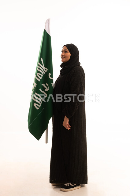 Pride in the Kingdom's achievements, the day we began, February 22, the anniversary of the founding of the first Saudi state in 1727 AD, showing loyalty and love for the homeland, a side portrait of a Saudi Arabian Gulf woman wearing a hijab and abaya holding the founding flag with gestures of pride and loftiness, full-length body, white background