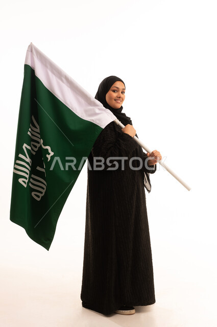 النظر إلى الكاميرا بإيماءات الاعتزاز والشموخ، ذكرى تأسيس الدولة السعودية الأولى 1727م، الفخر بإنجازات المملكة، يوم بدينا 22 فبراير، إظهار الولاء والحب للوطن, بورتريه لامرأة عربية خليجية سعودية ترتدي العباءة تحمل علم التأسيس، الجسم بالطول الكامل، خلفية بيضاء
