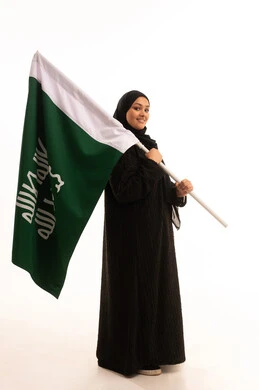 النظر إلى الكاميرا بإيماءات الاعتزاز والشموخ، ذكرى تأسيس الدولة السعودية الأولى 1727م، الفخر بإنجازات المملكة، يوم بدينا 22 فبراير، إظهار الولاء والحب للوطن, بورتريه لامرأة عربية خليجية سعودية ترتدي العباءة تحمل علم التأسيس، الجسم بالطول الكامل، خلفية بيضاء