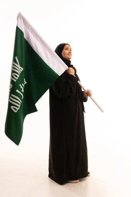 The day we began, February 22, the anniversary of the founding of the first Saudi state in 1727 AD, pride in the achievements of the Kingdom, showing loyalty and love for the homeland, a side portrait of a Saudi Arabian Gulf woman wearing a hijab and an abaya holding the founding flag with gestures of pride and loftiness, full-length body, white background