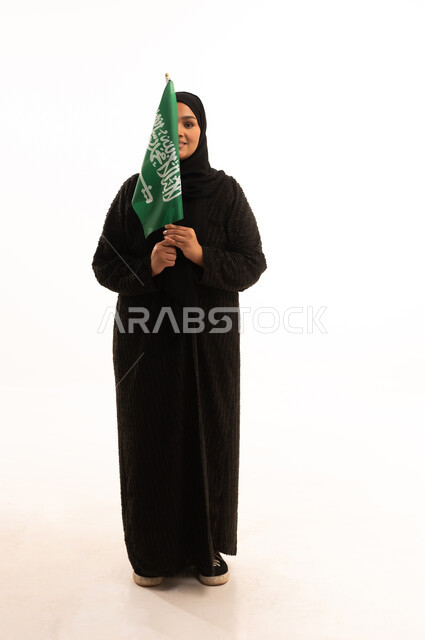 اليوم الوطني السعودي 23 سبتمبر، يوم العلم 11 مارس، مفهوم حب الوطن والانتماء اليه، بورتريه لسيدة عربية خليجية سعودية محجبة مبتسمة ترتدي عباءة سوداء تحمل بيدها علم المملكة وتنظر الى الكاميرا بإيماءات السرور والثقة بالنفس، صورة الجسم بالكامل، خلفية بيضاء