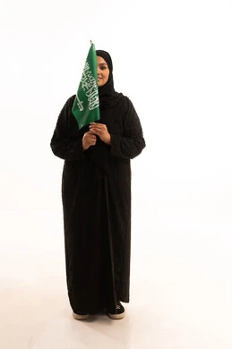 Saudi National Day September 23, Flag Day March 11, the concept of patriotism and belonging to the homeland, a portrait of a smiling veiled Saudi Arabian Gulf woman wearing a black abaya, holding the Kingdom's flag in her hand and looking at the camera with gestures of pleasure and self-confidence, a full body image, a white background