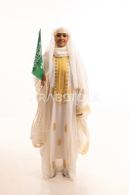 التباهي بإنجازات البلاد، النظر الى الكاميرا بتعابير الفخر، اليوم الوطني السعودي 23 سبتمبر، يوم العلم 11 مارس، بورتريه لامرأة عربية خليجية سعودية مبتسمة ترتدي الشيلة والنشل تحمل بيدها علم المملكة، ارتداء أزياء المنطقة الشرقية، الجسم الكامل بالطول، خلفية بيضاء