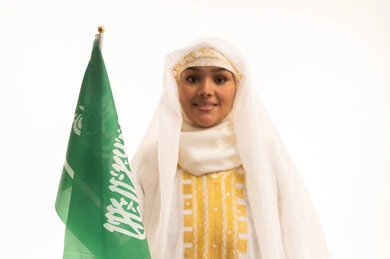 الاعتزاز بالانتماء للوطن، التباهي بإنجازات البلاد، النظر الى الكاميرا بتعابير الفخر، اليوم الوطني السعودي 23 سبتمبر، يوم العلم 11 مارس، بورتريه مقرب لامرأة عربية خليجية سعودية مبتسمة ترتدي الشيلة والنشل تحمل بيدها علم المملكة، ارتداء أزياء المنطقة الشرقية، خلفية بيضاء