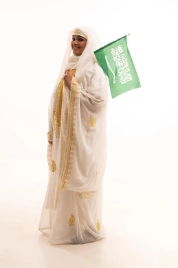 ارتداء أزياء المنطقة الشرقية، التباهي بإنجازات البلاد، النظر الى الكاميرا بتعابير الفخر، اليوم الوطني السعودي 23 سبتمبر، يوم العلم 11 مارس، بورتريه لامرأة عربية خليجية سعودية مبتسمة ترتدي الشيلة والنشل تحمل بيدها علم المملكة، الجسم الكامل بالطول، خلفية بيضاء