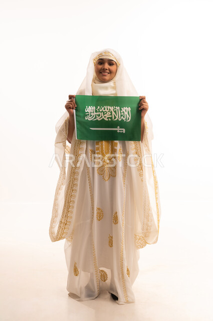 Boasting about the country's achievements, looking at the camera with an expression of pride, Saudi National Day September 23, Flag Day March 11, portrait of a smiling Saudi Arabian Gulf woman wearing a sheila and a neshel holding the flag of the Kingdom, wearing the Eastern Province costumes, full body length, white background