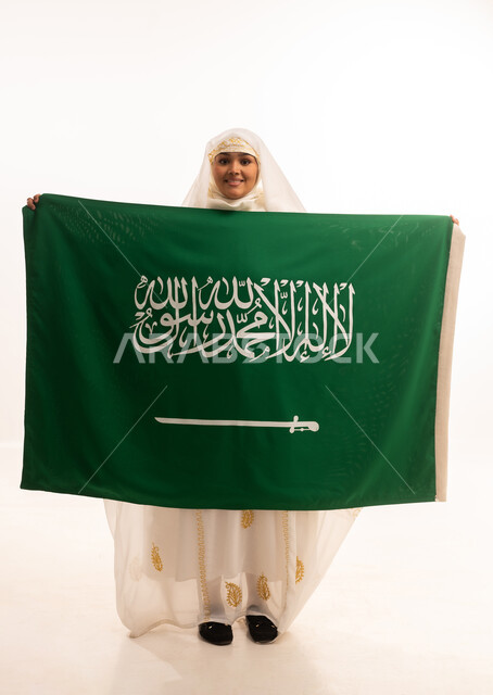 التباهي بإنجازات البلاد، النظر الى الكاميرا بتعابير الفخر، اليوم الوطني السعودي 23 سبتمبر، يوم العلم 11 مارس، بورتريه لامرأة عربية خليجية سعودية مبتسمة ترتدي الشيلة والنشل تحمل بيدها علم المملكة، ارتداء أزياء المنطقة الشرقية، الجسم الكامل بالطول، خلفية بيضاء