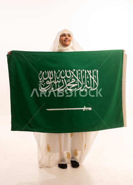التطلع إلى الأعلى بإيماءات الشموخ، ارتداء أزياء المنطقة الشرقية، التباهي بإنجازات البلاد، اليوم الوطني السعودي 23 سبتمبر، يوم العلم 11 مارس، بورتريه لامرأة عربية خليجية سعودية مبتسمة ترتدي الشيلة والنشل تحمل بيدها علم المملكة، الجسم الكامل بالطول، خلفية بيضاء