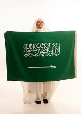 التطلع إلى الأعلى بإيماءات الشموخ، ارتداء أزياء المنطقة الشرقية، التباهي بإنجازات البلاد، اليوم الوطني السعودي 23 سبتمبر، يوم العلم 11 مارس، بورتريه لامرأة عربية خليجية سعودية مبتسمة ترتدي الشيلة والنشل تحمل بيدها علم المملكة، الجسم الكامل بالطول، خلفية بيضاء