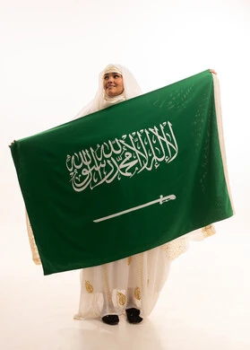 النظر إلى الأعلى بإيماءات الشموخ، ارتداء أزياء المنطقة الشرقية، التباهي بإنجازات البلاد، اليوم الوطني السعودي 23 سبتمبر، يوم العلم 11 مارس، بورتريه لامرأة عربية خليجية سعودية مبتسمة ترتدي الشيلة والنشل تحمل بيدها علم المملكة، الجسم الكامل بالطول، خلفية بيضاء