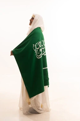 إيماءات الشموخ والفخر، ارتداء أزياء المنطقة الشرقية، التباهي بإنجازات البلاد، اليوم الوطني السعودي 23 سبتمبر، يوم العلم 11 مارس، بورتريه من الجانب لامرأة عربية خليجية سعودية مبتسمة ترتدي الشيلة والنشل تضع على كتفيها علم المملكة، الجسم الكامل بالطول، خلفية بيضاء