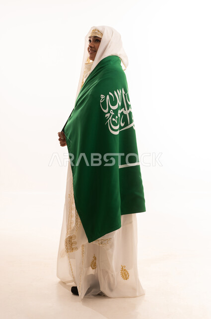 إيماءات الشموخ والفخر، ارتداء أزياء المنطقة الشرقية، التباهي بإنجازات البلاد، اليوم الوطني السعودي 23 سبتمبر، يوم العلم 11 مارس، بورتريه من الجانب لامرأة عربية خليجية سعودية مبتسمة ترتدي الشيلة والنشل تضع على كتفيها علم المملكة، الجسم الكامل بالطول، خلفية بيضاء