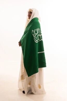 Gestures of pride and dignity, wearing the Eastern Province costumes, bragging about the country's achievements, Saudi National Day September 23, Flag Day March 11, side portrait of a smiling Saudi Arabian Gulf Arab woman wearing a sheyla and a neshel, placing the Kingdom's flag on her shoulders, full body length, white background