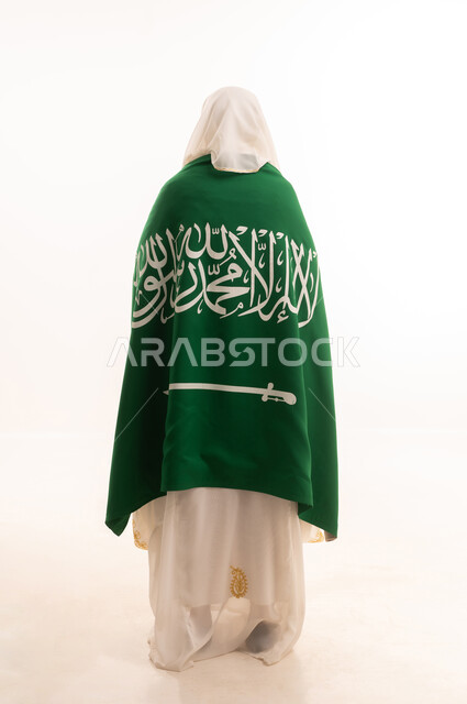 اليوم الوطني السعودي 23 سبتمبر، ارتداء أزياء المنطقة الشرقية، التباهي بإنجازات البلاد، يوم العلم 11 مارس، بورتريه من الخلف لامرأة عربية خليجية سعودية ترتدي الشيلة والنشل تضع على كتفيها علم المملكة، الجسم الكامل بالطول، خلفية بيضاء