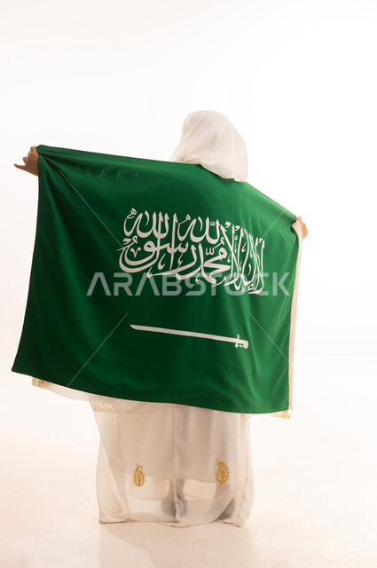 الاحتفال باليوم الوطني السعودي 23 سبتمبر، ارتداء أزياء المنطقة الشرقية، التباهي بإنجازات البلاد، يوم العلم 11 مارس، بورتريه من الخلف لامرأة عربية خليجية سعودية ترتدي الشيلة والنشل تضع على كتفيها علم المملكة، الجسم الكامل بالطول، خلفية بيضاء