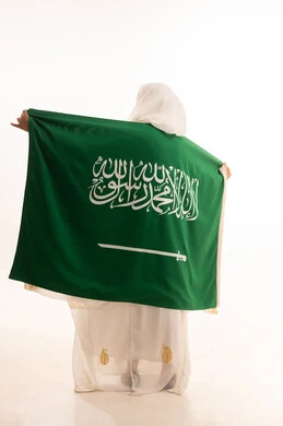 الاحتفال باليوم الوطني السعودي 23 سبتمبر، ارتداء أزياء المنطقة الشرقية، التباهي بإنجازات البلاد، يوم العلم 11 مارس، بورتريه من الخلف لامرأة عربية خليجية سعودية ترتدي الشيلة والنشل تضع على كتفيها علم المملكة، الجسم الكامل بالطول، خلفية بيضاء
