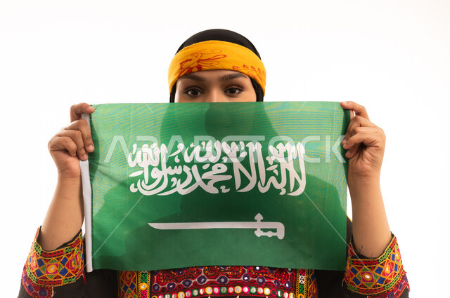 الاحتفال بالمناسبات الوطنية، مفهوم حب الوطن، اليوم الوطني السعودي 23 سبتمبر, يوم بدينا 1727م, الفخر بالتراث التاريخي، يوم العلم 11 مارس، ارتداء زي المنطقة الوسطى، بورتريه مقرب لإمرأة عربية خليجية سعودية ترتدي الكرتة والهامة تحمل علم المملكة, خلفية بيضاء