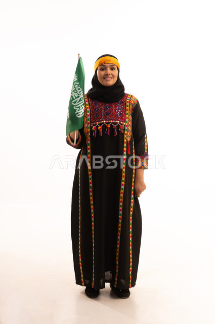 الفخر بالتراث التاريخي، يوم العلم 11 مارس، يوم بدينا 1727م, ارتداء زي المنطقة الوسطى، اليوم الوطني السعودي 23 سبتمبر, بورتريه لإمرأة عربية خليجية سعودية ترتدي الكرتة والهامة تحمل علم المملكة تنظر إلى الكاميرا بتعابير الفخر, الجسم بالطول الكامل، خلفية بيضاء