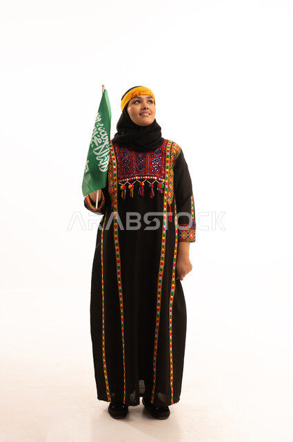 يوم بدينا 1727م, الاعتزاز والتباهي بإنجازات البلاد، الفخر بالتراث التاريخي، يوم العلم 11 مارس، ارتداء زي المنطقة الوسطى، اليوم الوطني السعودي 23 سبتمبر, بورتريه لإمرأة عربية خليجية سعودية ترتدي الكرتة والهامة تحمل علم المملكة, الجسم بالطول الكامل، خلفية بيضاء