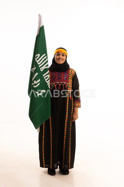 ذكرى تأسيس الدولة السعودية الأولى 22 فبراير، يوم بدينا 1727 م، ارتداء أزياء المنطقة الوسطى، بورتريه لإمرأة عربية خليجية سعودية مبتسمة ترتدي الكرتة والهامة تحمل بيدها علم يوم التأسيس، الاحتفال بالمناسبات والأعياد الوطنية الشعبية، الجسم بالطول الكامل، خلفية بيضاء